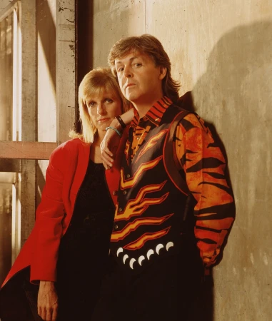 Portrait of adult male and female wearing 1980s fashion, posing against a concrete wall with metal framing in the background.