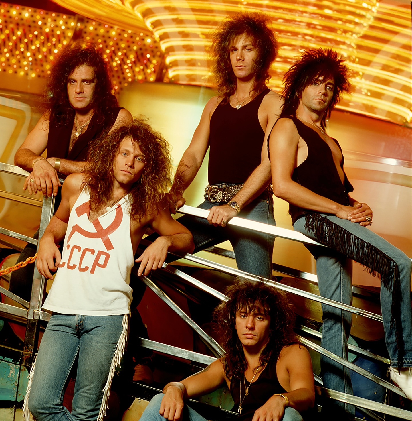 Group of five men with long hair in tank tops and jeans pose on a staircase in front of a brightly lit background, in a studio.