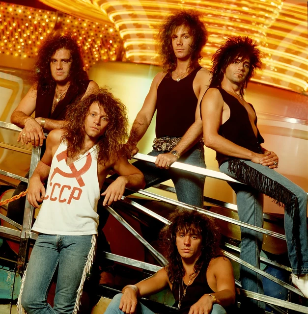 Group of five men with long hair in tank tops and jeans pose on a staircase in front of a brightly lit background, in a studio.