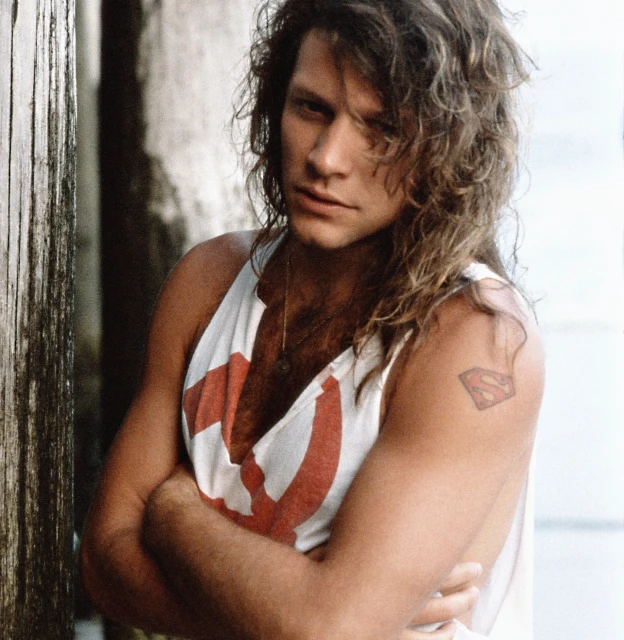 Jon Bon Jovi gazes with arms crossed, wearing a white tank top with a red design, in a close-up shot against a neutral background.