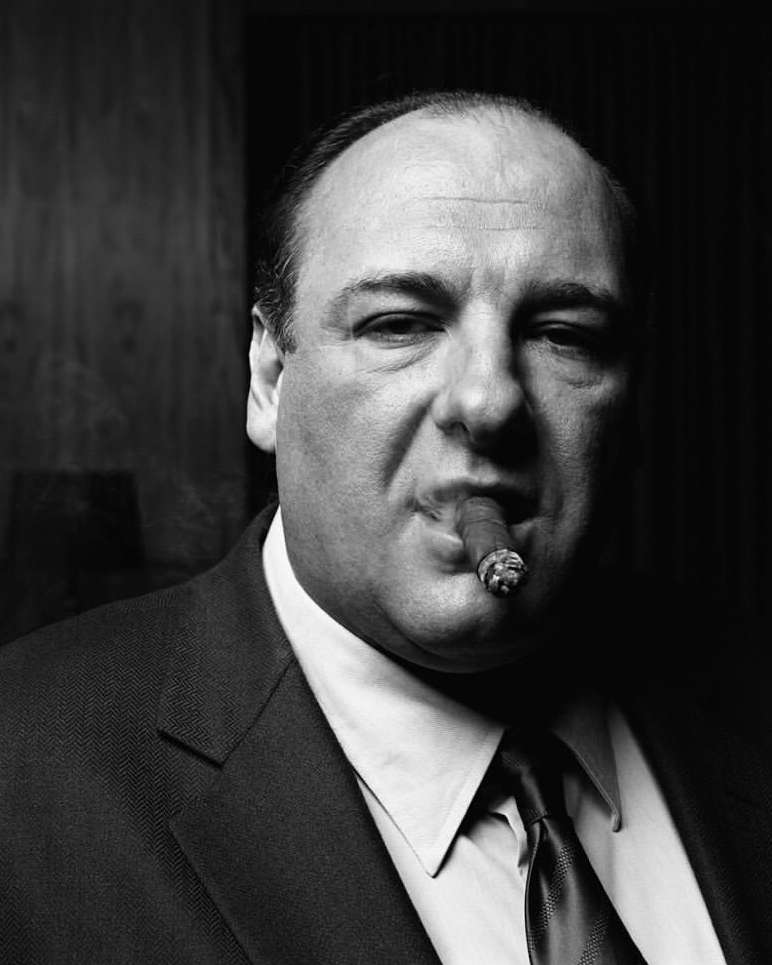 Monochrome portrait of an adult male smoking a cigar, wearing a dark suit and tie, set against a dark background, photographed indoors.