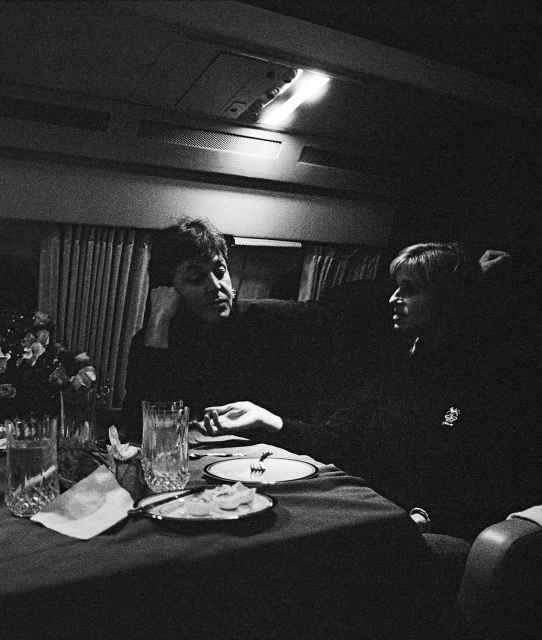 Two adults seated at a formal table with crystal glasses, flowers, plates, silverware, and overhead lighting in a dark indoor space.