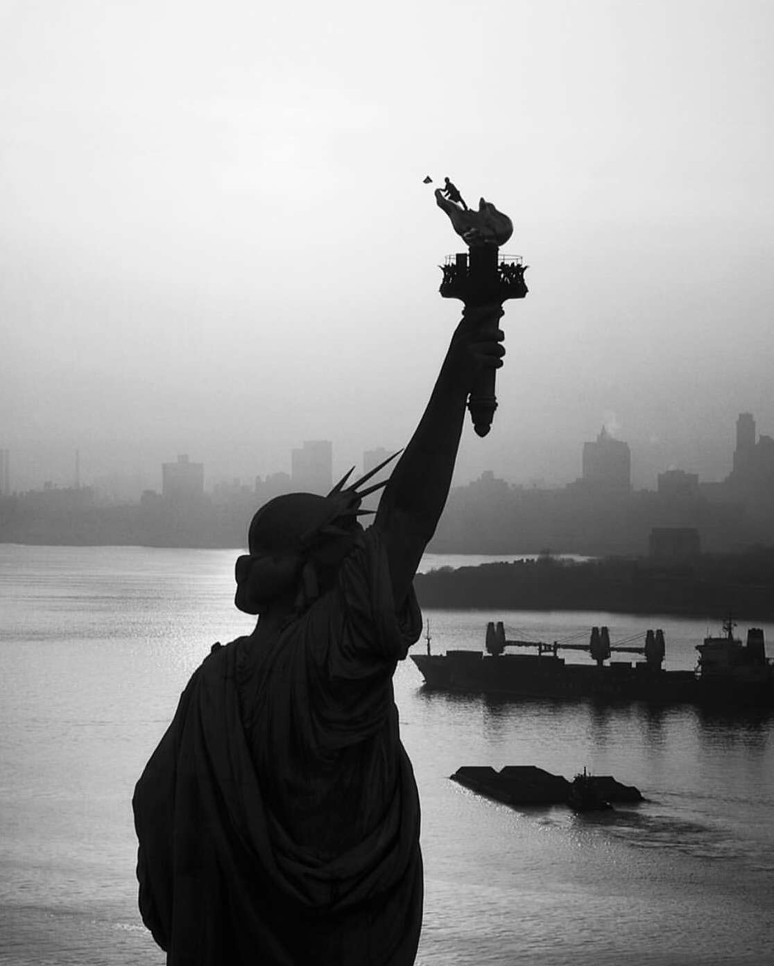 Statue of Liberty at dusk, silhouetted against the city skyline and waterfront in black and white, featuring a ship in the background.