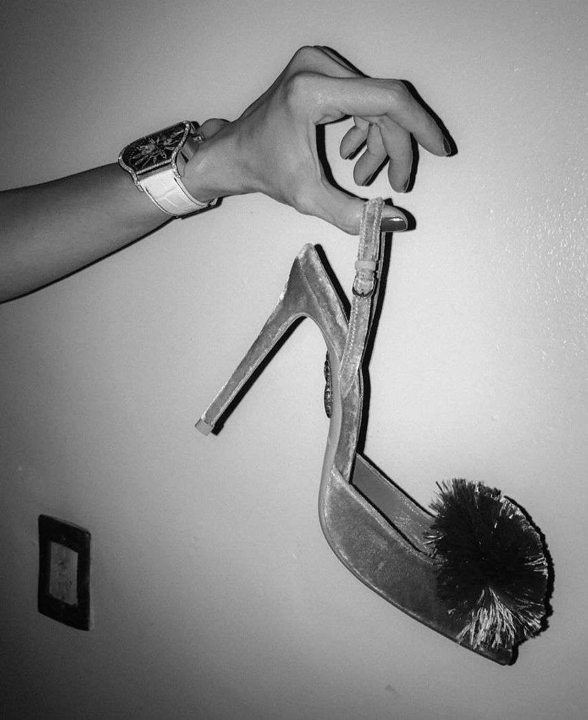 A woman's hand holds a high heel with a fluffy pompom, wrist adorned with a stylish watch, against a neutral wall backdrop in a studio setting.