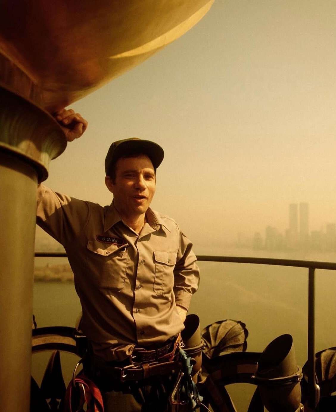 Man in a cap and uniform standing next to a metal structure with a cityscape featuring the Twin Towers in the background, warm light.