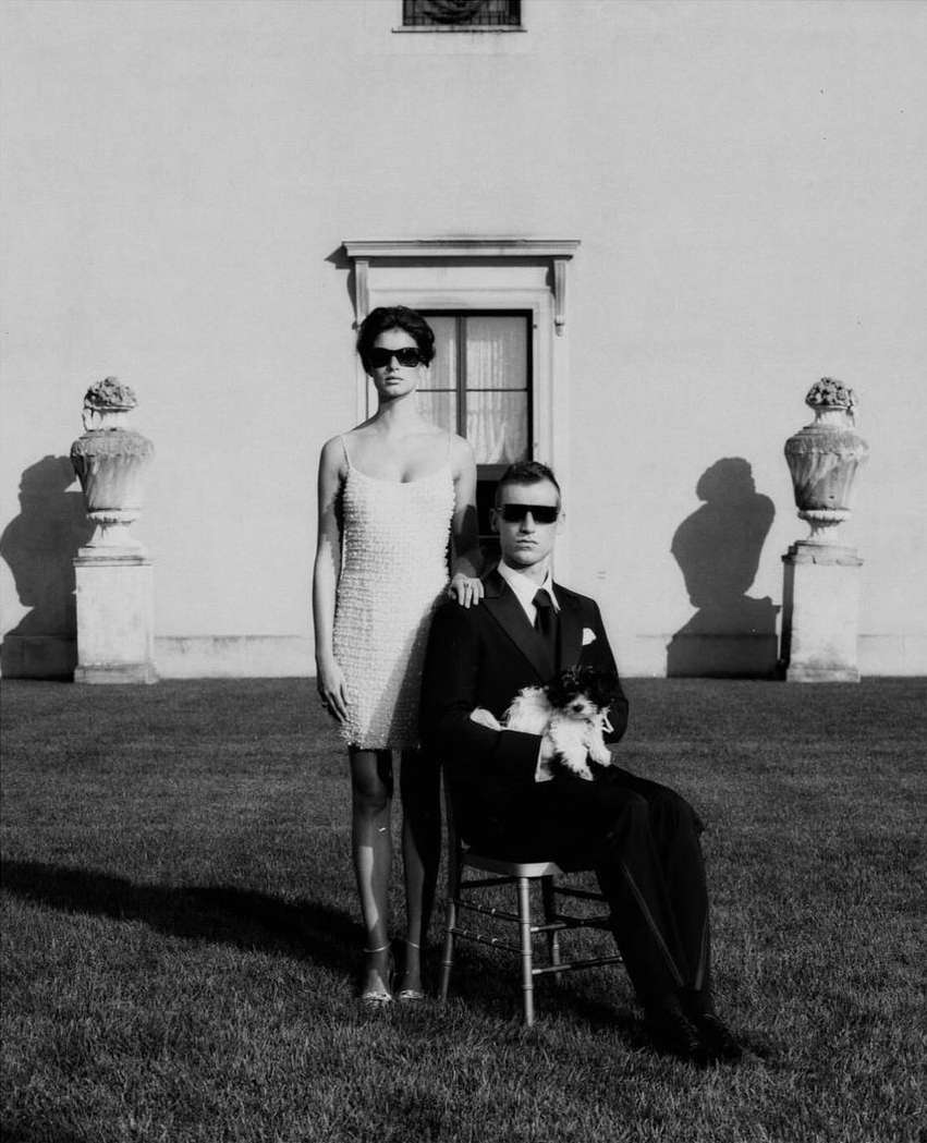 Black-and-white outdoor shot of a formally dressed couple standing on grass with a dog. Man seated, woman standing, sunglasses, white building.