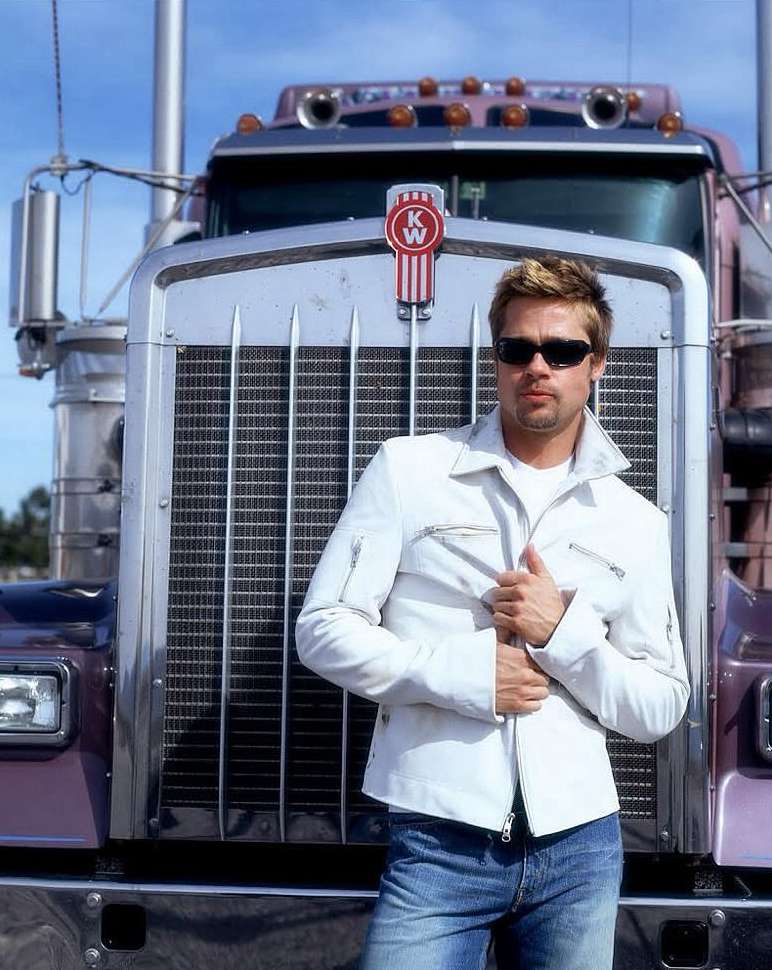 Adult male in sunglasses poses in front of a semi-truck wearing a white jacket and blue jeans outdoors during daylight in portrait orientation.