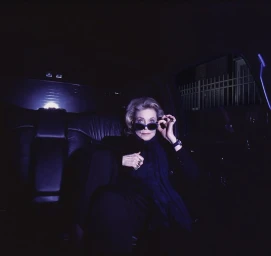 An elderly woman wearing dark sunglasses sitting inside a dark car interior, photographed by Timothy White.