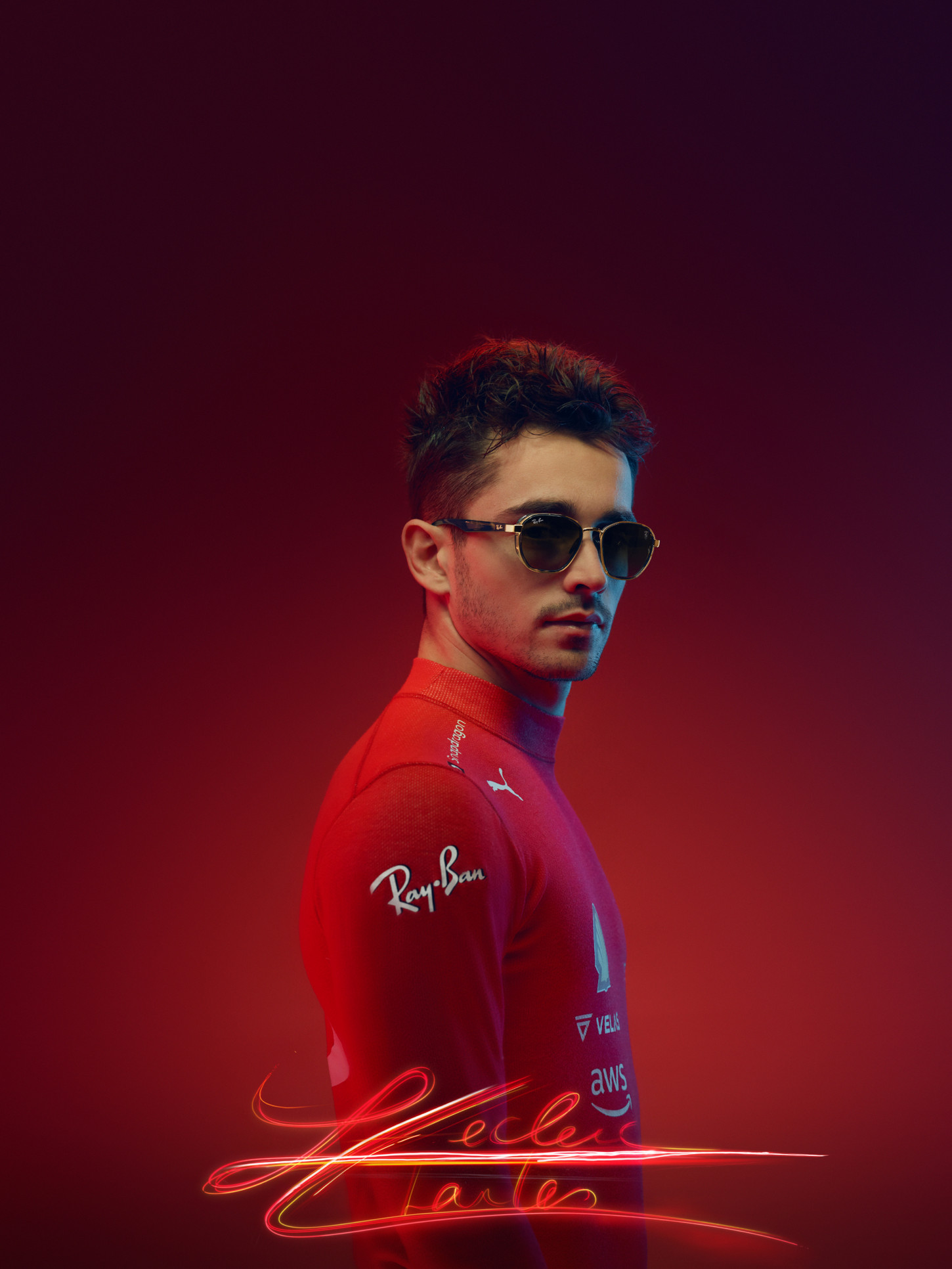 Portrait of a young adult male wearing a red racing suit and sunglasses, against a deep red studio backdrop with soft lighting.