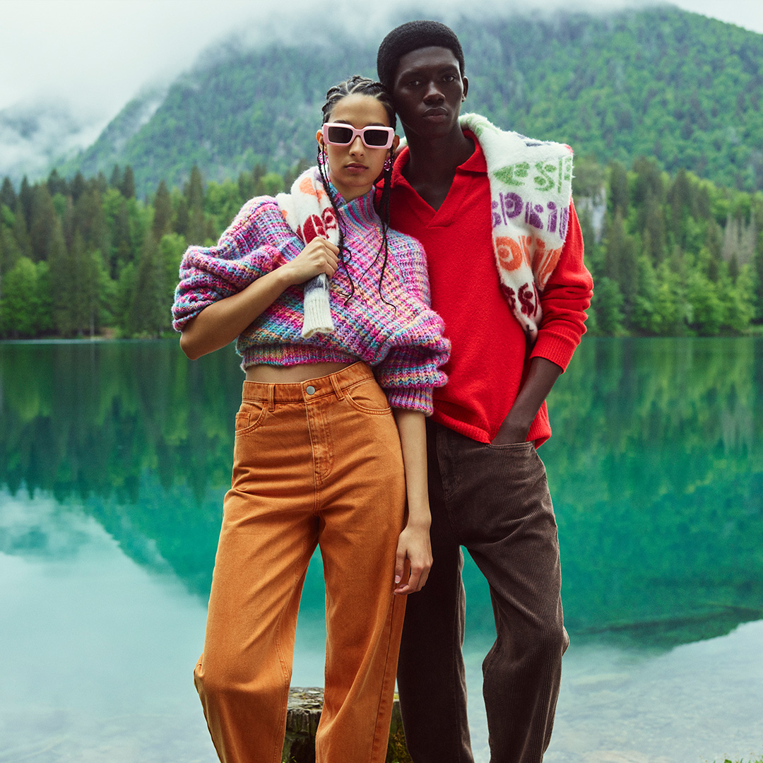 Two fashion models, one adult woman and one adult man, pose outdoors near a lake with mountains in the background wearing casual clothing.