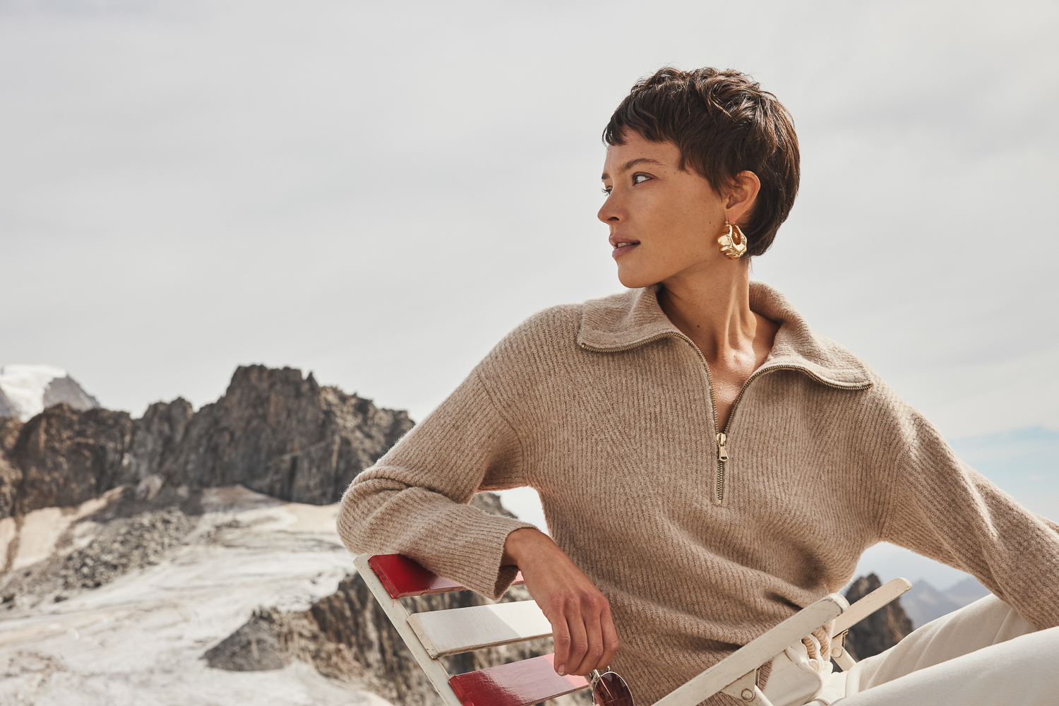 Adult female with short brown hair sits on a chair, wearing a beige sweater and white pants, posing outdoors with a mountain backdrop.