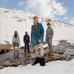 Four models in winter apparel standing in a snowy mountain landscape near a rocky stream under a clear sky.