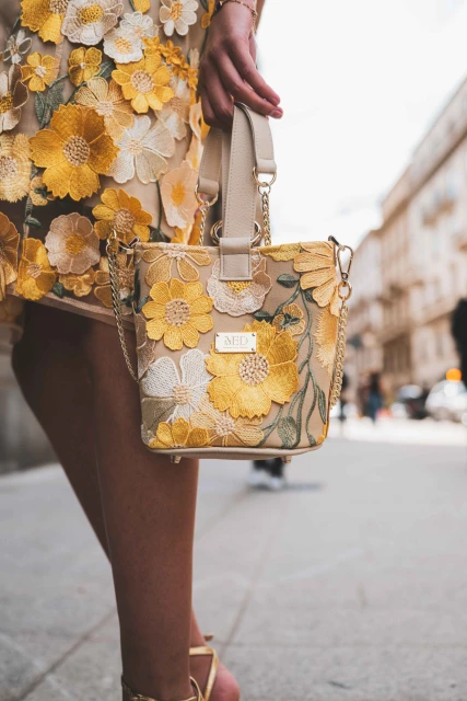 Woman carrying a floral embroidered handbag in an urban setting, complementing her flower-adorned dress, with a sunny and bright ambiance.