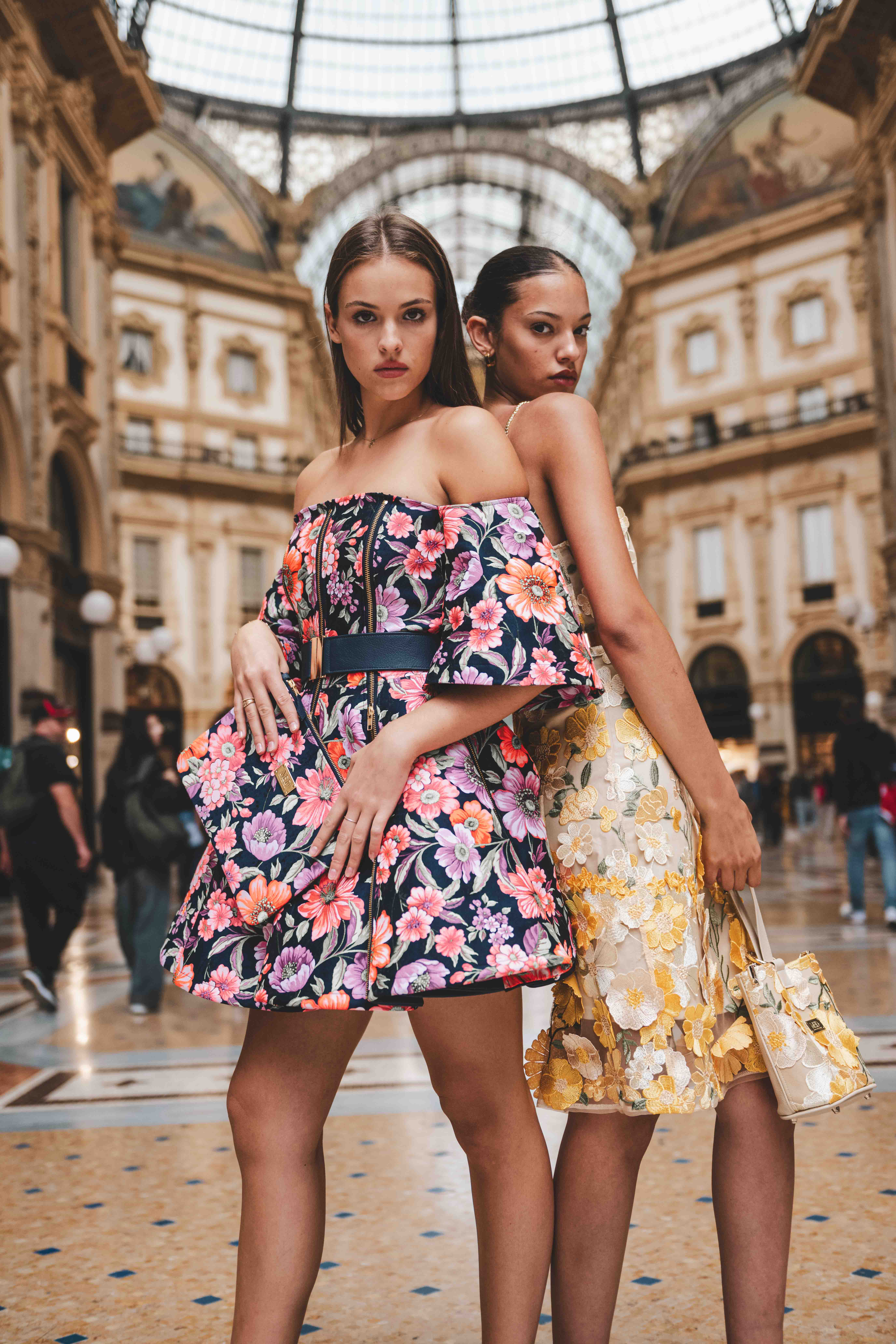 Two women pose in vibrant floral dresses inside Galleria Vittorio Emanuele II, Milan, Italy. High fashion scene with architectural backdrop.