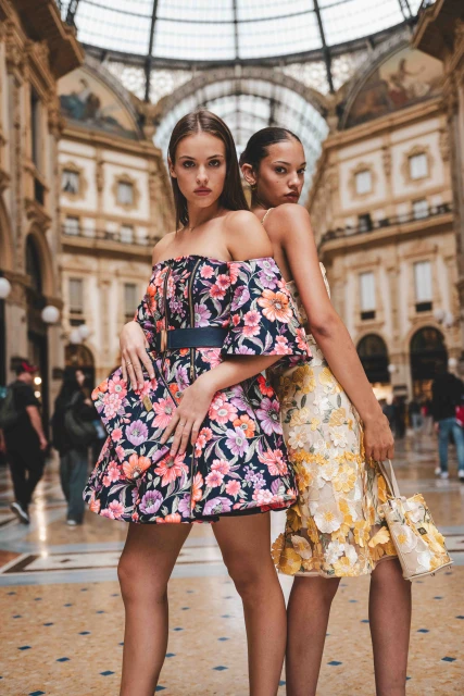 Two women pose in vibrant floral dresses inside Galleria Vittorio Emanuele II, Milan, Italy. High fashion scene with architectural backdrop.
