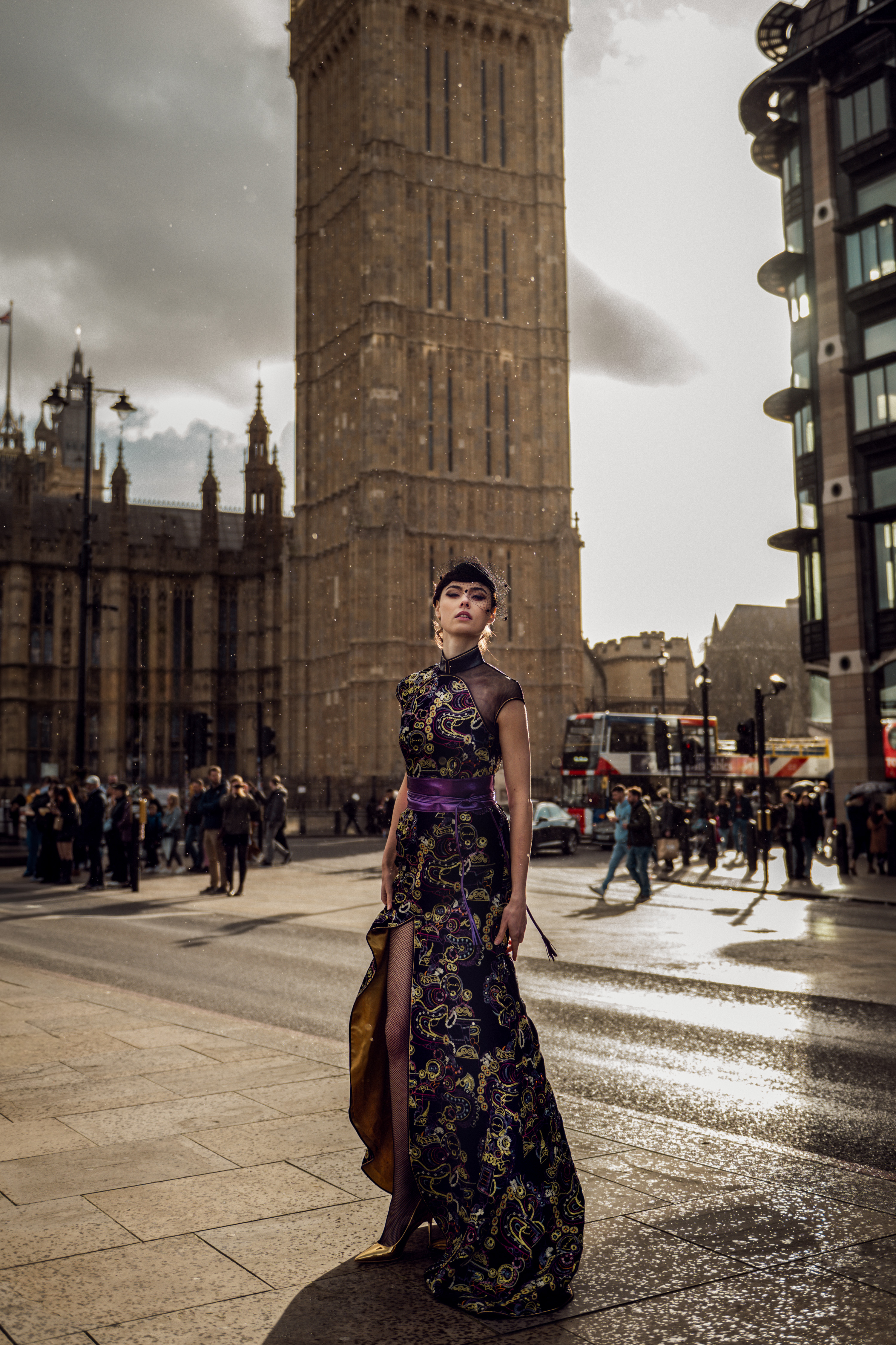 Fashionable woman in ornate gown poses in city street, London landmark in background, pedestrians and traffic create urban scene.