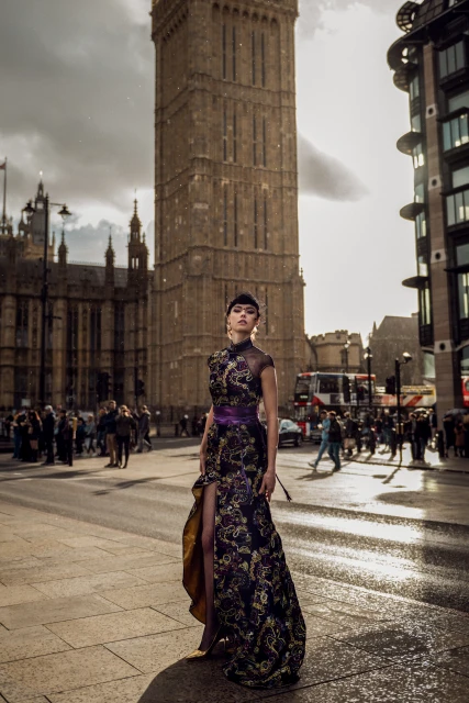 Fashionable woman in ornate gown poses in city street, London landmark in background, pedestrians and traffic create urban scene.