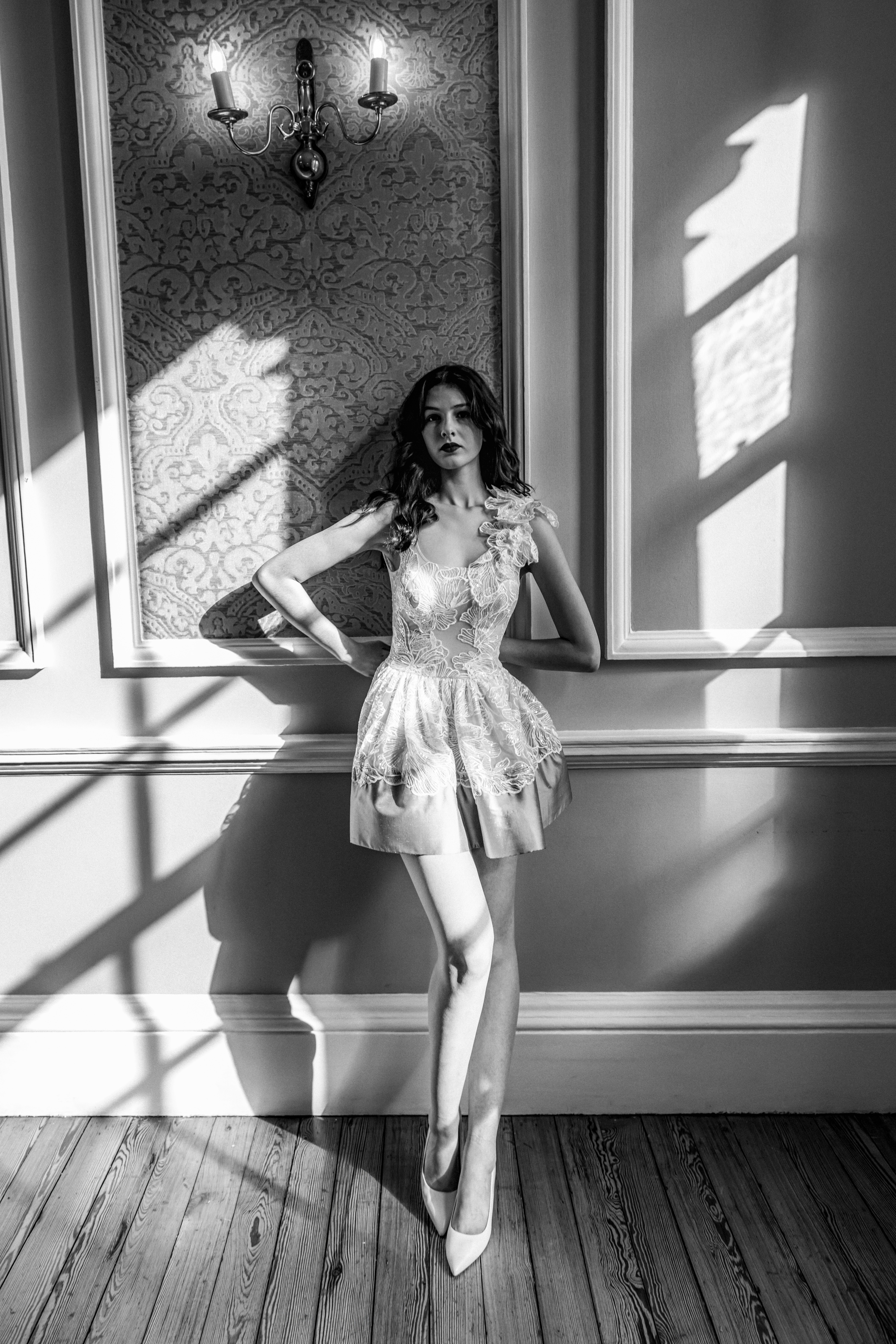 Elegant woman in a short dress posing in a room with paneled walls and patterned wallpaper, bathed in soft window light, black and white.