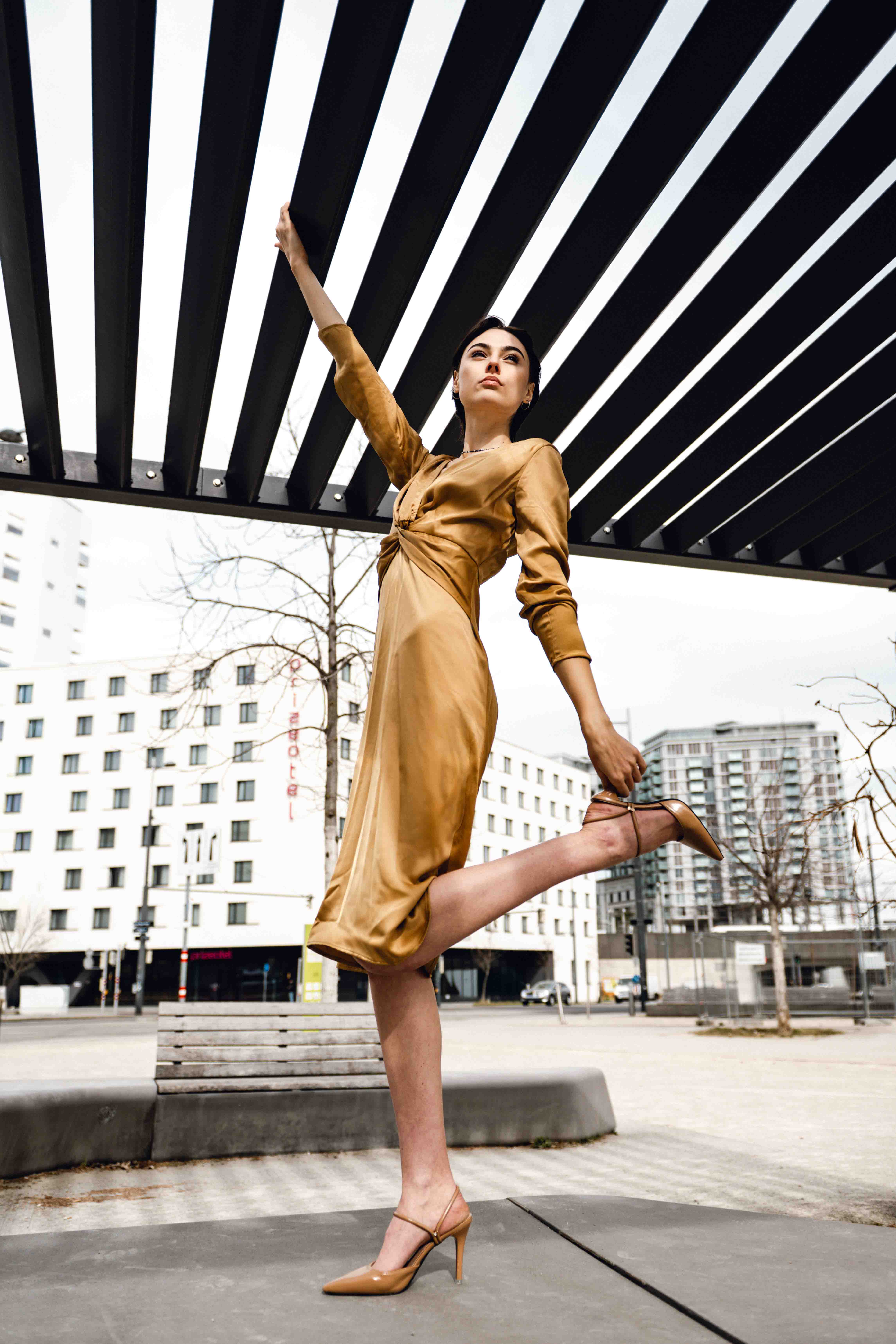 An adult female fashion model posing in an outdoor urban setting, wearing a gold dress and strappy high-heeled shoes near modern buildings.