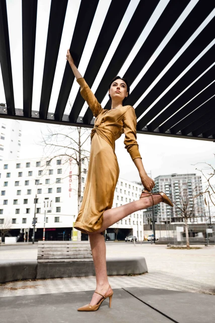 An adult female fashion model posing in an outdoor urban setting, wearing a gold dress and strappy high-heeled shoes near modern buildings.
