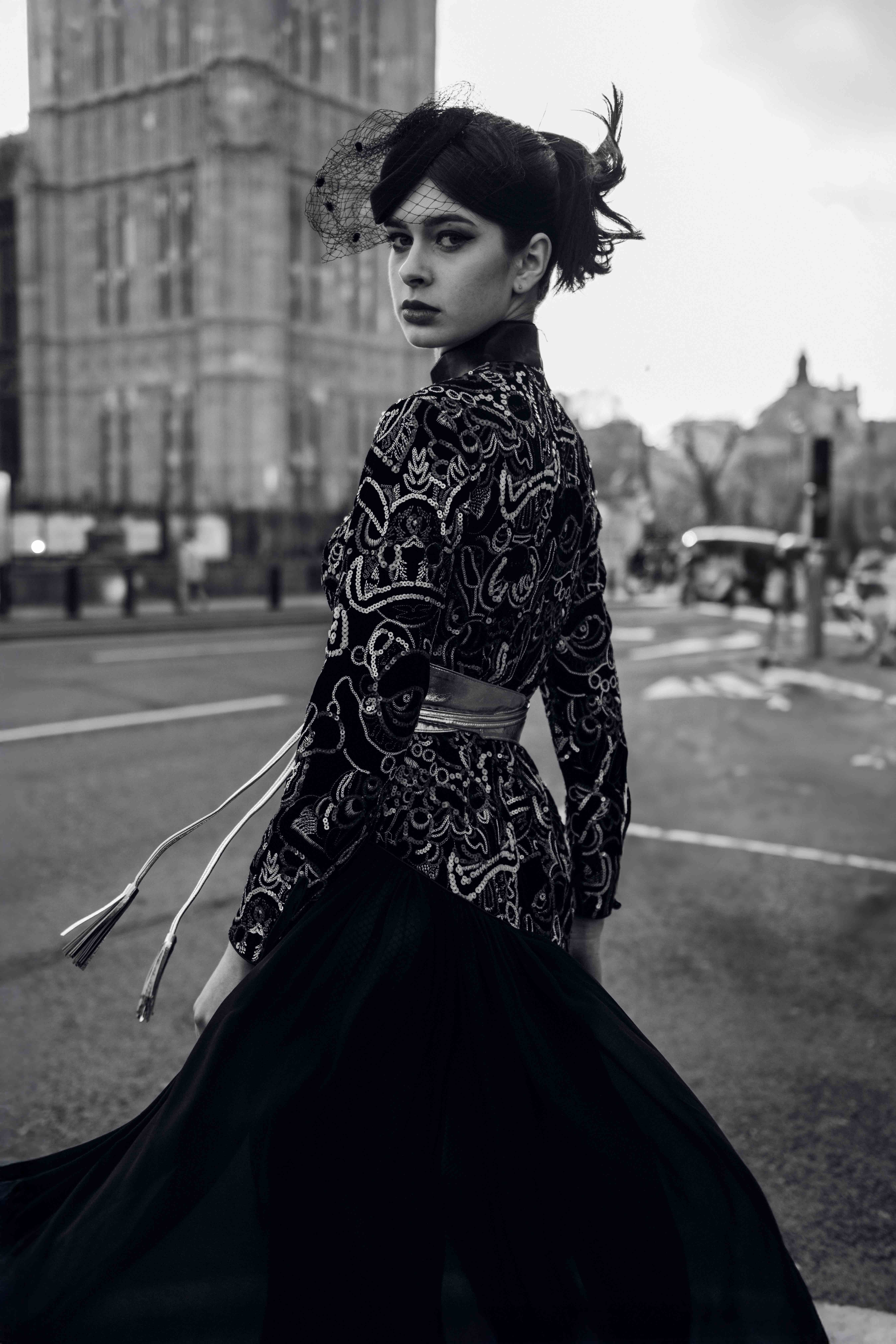 Black and white portrait of a young woman in patterned jacket and flowing skirt posing on a street with a large building in the background.