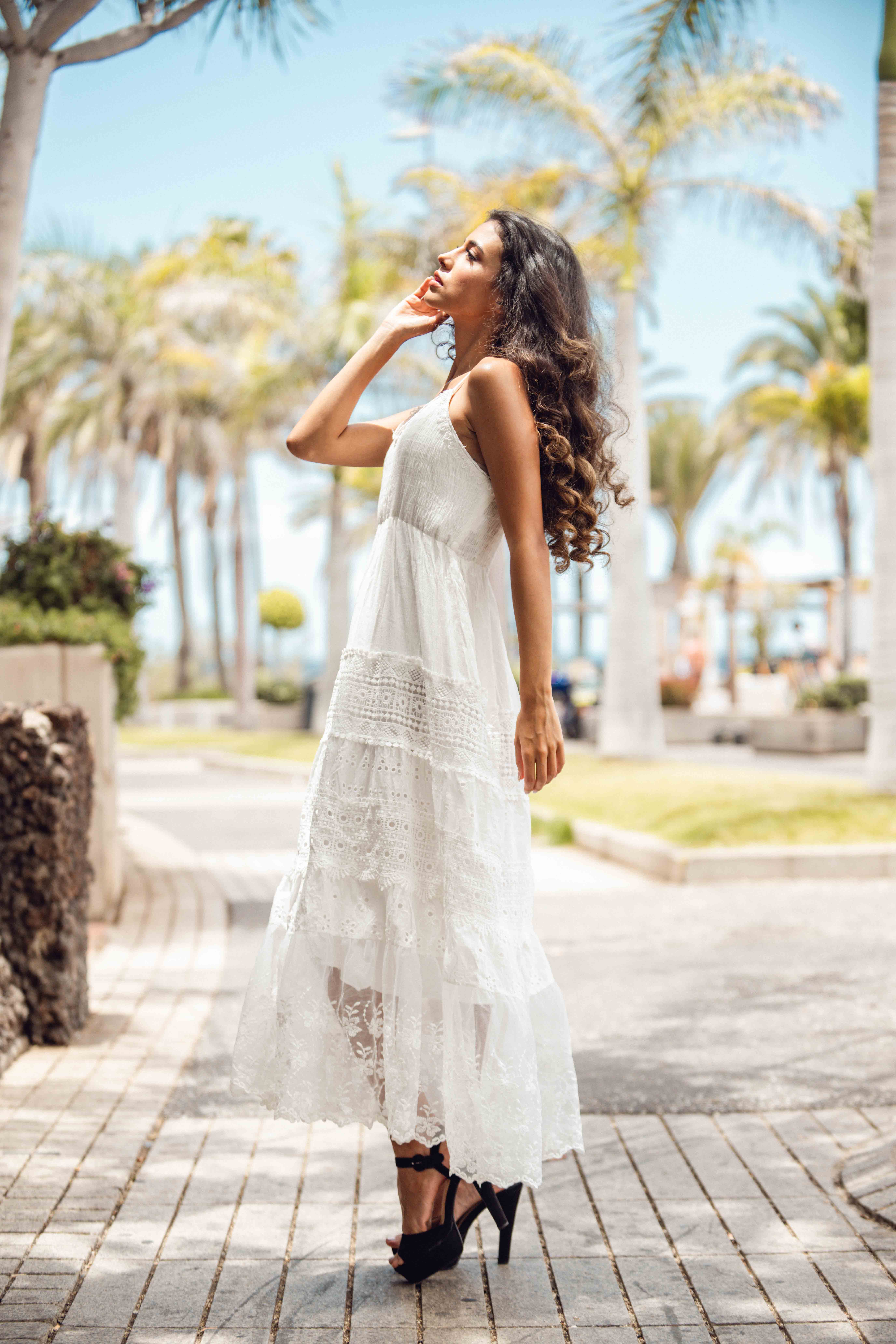 Woman in a white lace dress stands on a paved path lined with palm trees under a clear blue sky in an outdoor setting.