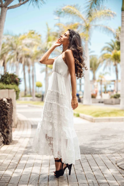 Woman in a white lace dress stands on a paved path lined with palm trees under a clear blue sky in an outdoor setting.