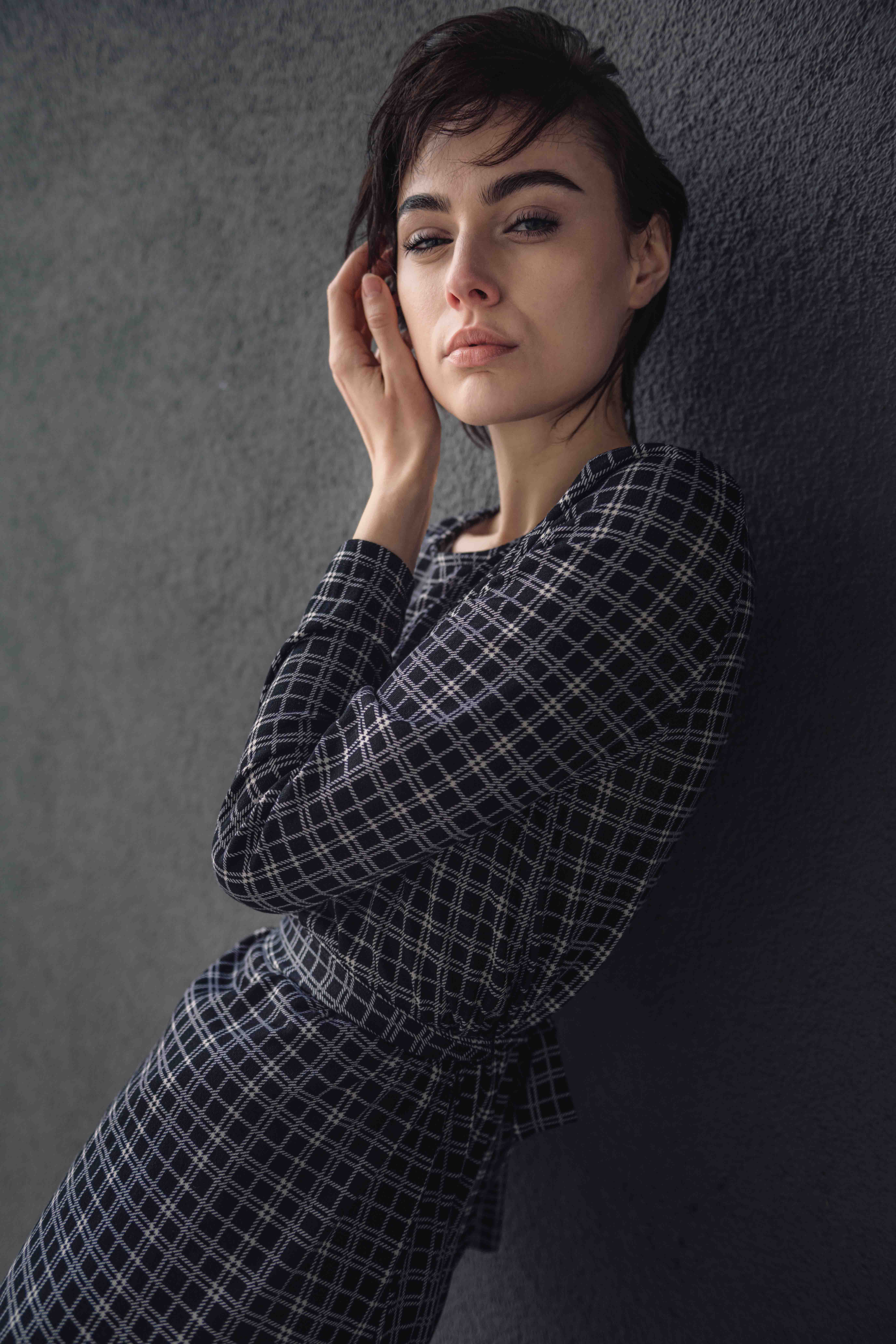 Portrait of a young woman with short dark hair, wearing a checkered dress, posing against a textured grey wall in soft, diffused lighting.