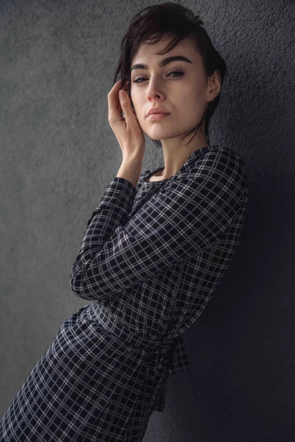 Portrait of a young woman with short dark hair, wearing a checkered dress, posing against a textured grey wall in soft, diffused lighting.