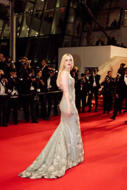 Elle Fanning wearing a sequined gown on a red carpet, posing and looking back at the camera with photographers in the background.