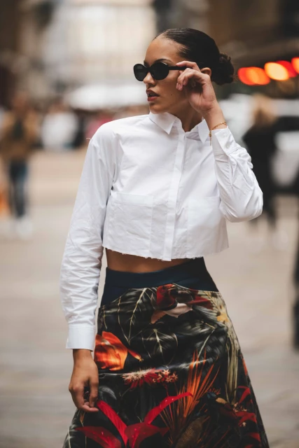 Fashionable woman in sunglasses, cropped white shirt, and floral skirt walks outdoors against a blurred urban background on a sunny day.