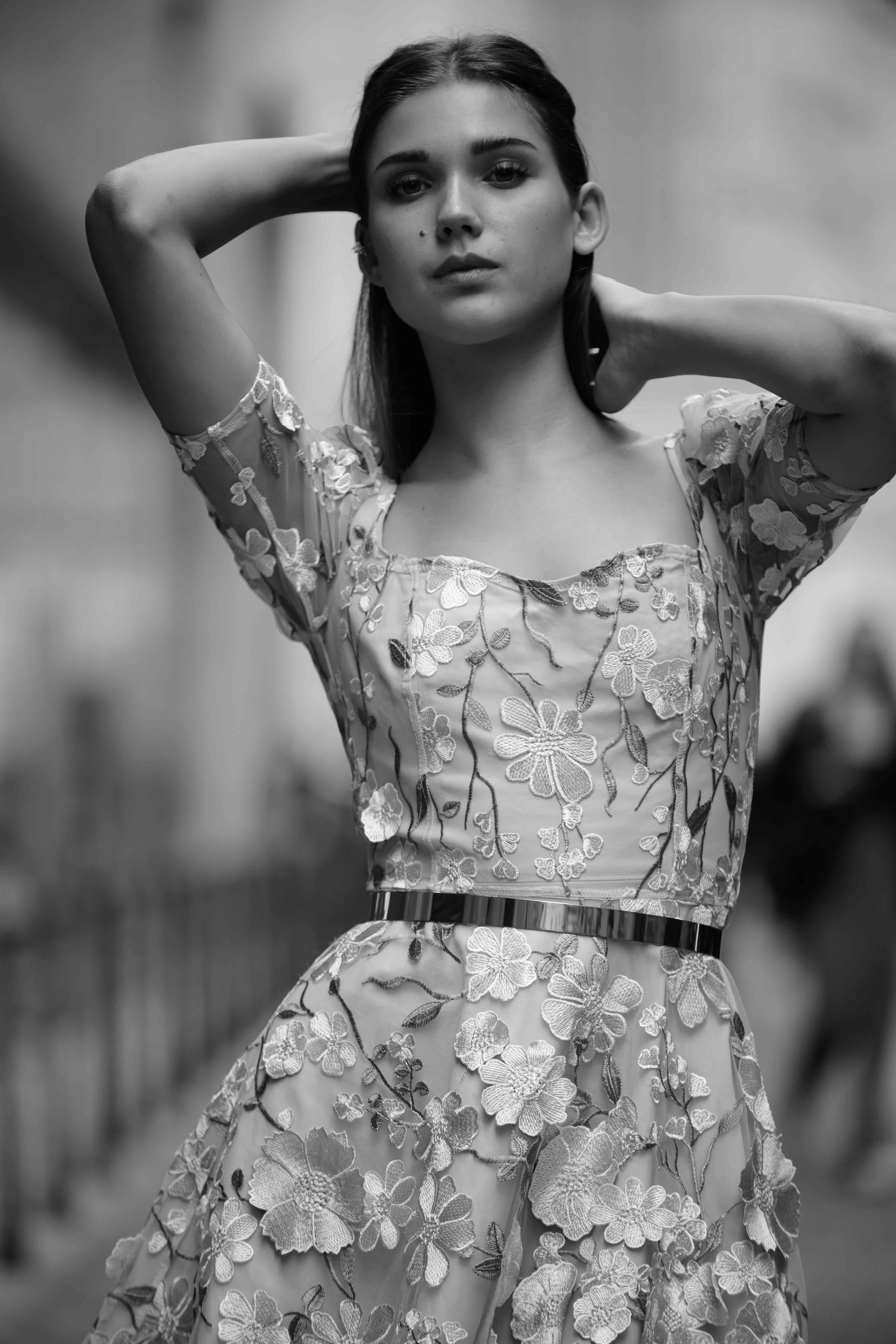 Monochrome portrait of a young woman in a floral dress with hands behind her head, showcasing fashion and style in an urban setting.