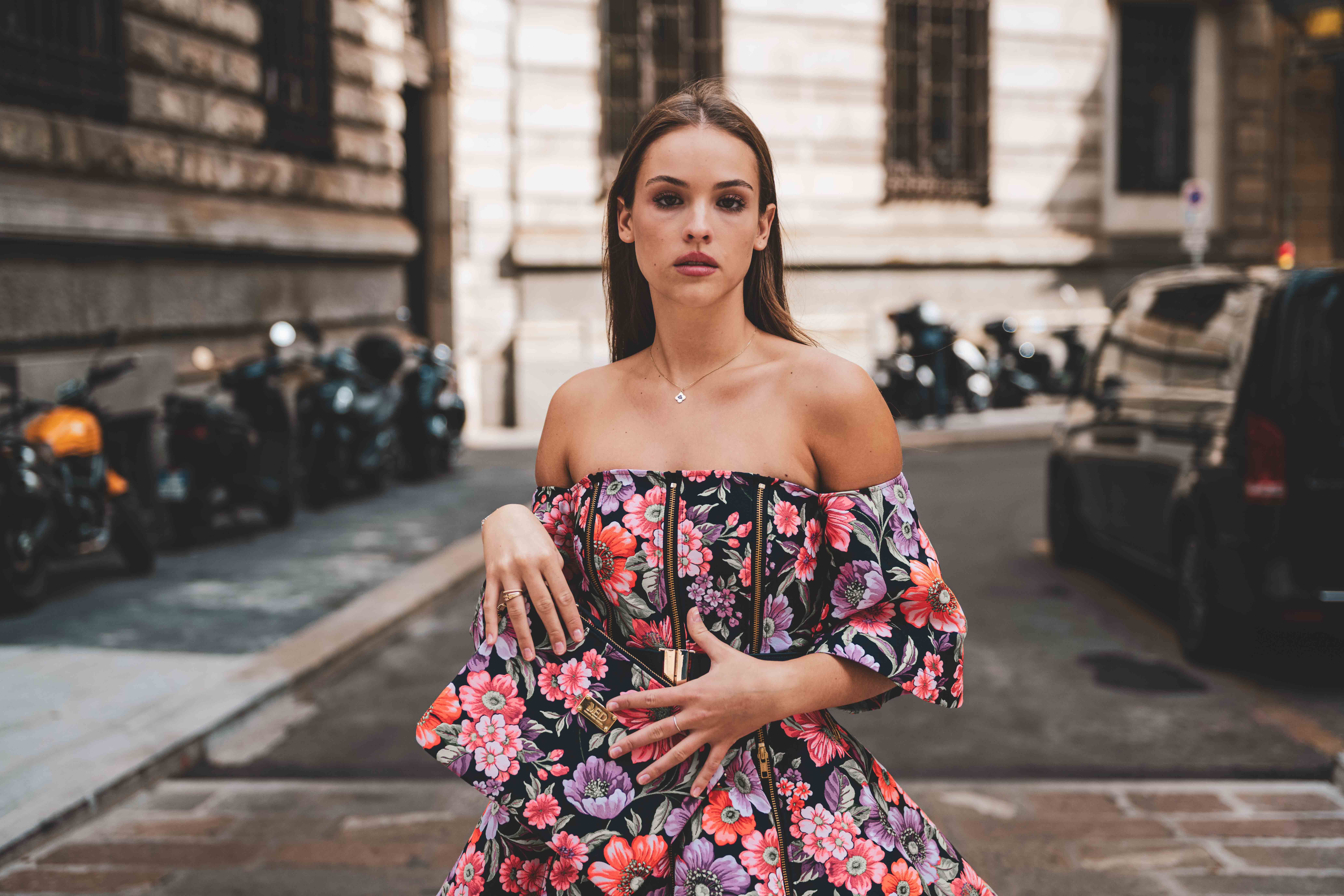 Brunette adult woman in floral dress with clutch poses on city street with parked motorbikes and black car in the background.