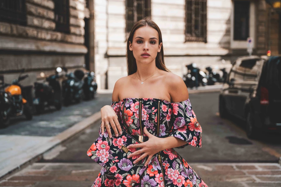 Brunette adult woman in floral dress with clutch poses on city street with parked motorbikes and black car in the background.