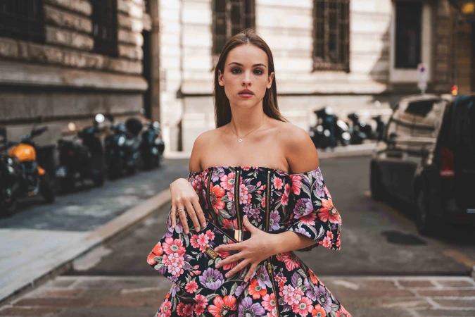 Brunette adult woman in floral dress with clutch poses on city street with parked motorbikes and black car in the background.