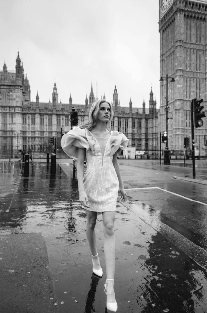 Monochrome fashion portrait of blonde woman in a short dress with puffy sleeves, walking in a city with a large clock tower background.