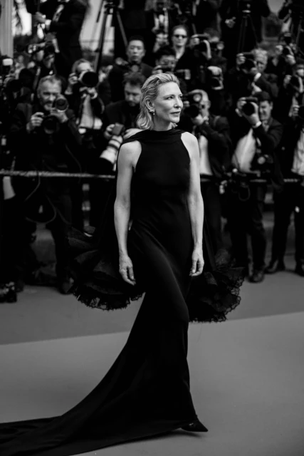 Cate Blanchett on the red carpet wearing a black halter gown with a long train, surrounded by photographers during a red carpet event.