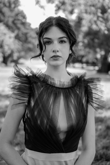 Headshot of a young woman with dark hair wearing a black tulle top standing in a grassy outdoor setting with blurred trees in background.