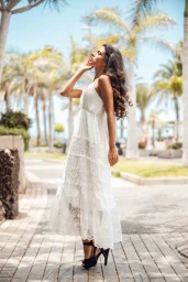 Woman with long brunette hair posing in a tiered white lace maxi dress and black high heels on a palm tree lined pathway.
