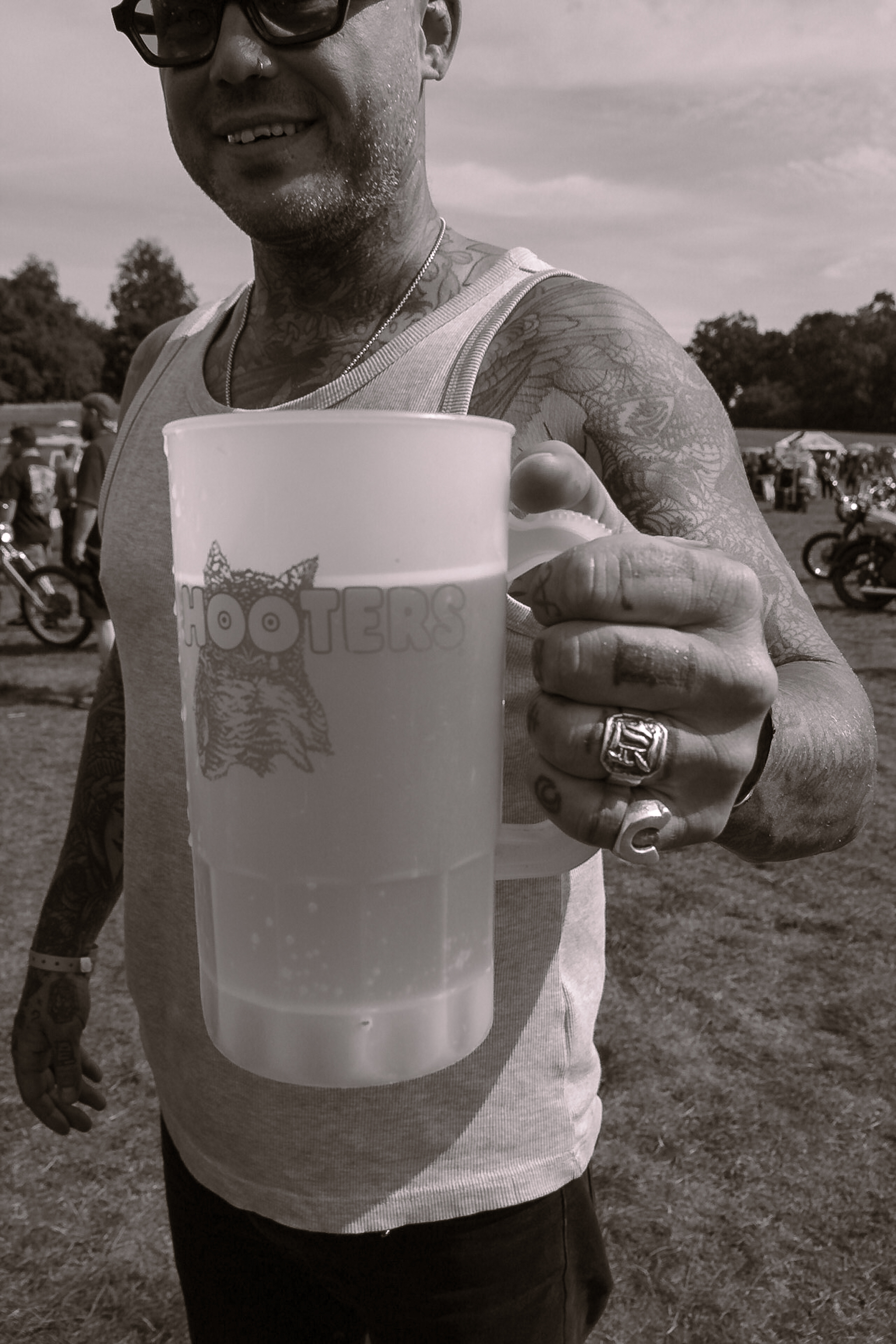 Man holding Hooters beer mug outdoors in monochrome. Tattooed arm, rings, sunglasses, tank top, and background with motorbikes and trees.