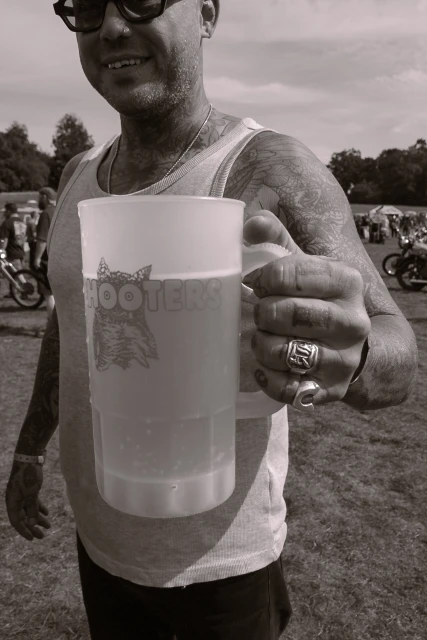 Man holding Hooters beer mug outdoors in monochrome. Tattooed arm, rings, sunglasses, tank top, and background with motorbikes and trees.