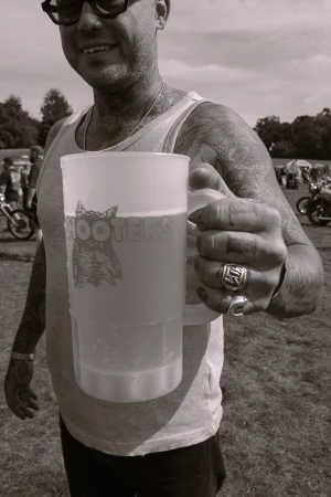 Man holding Hooters beer mug outdoors in monochrome. Tattooed arm, rings, sunglasses, tank top, and background with motorbikes and trees.