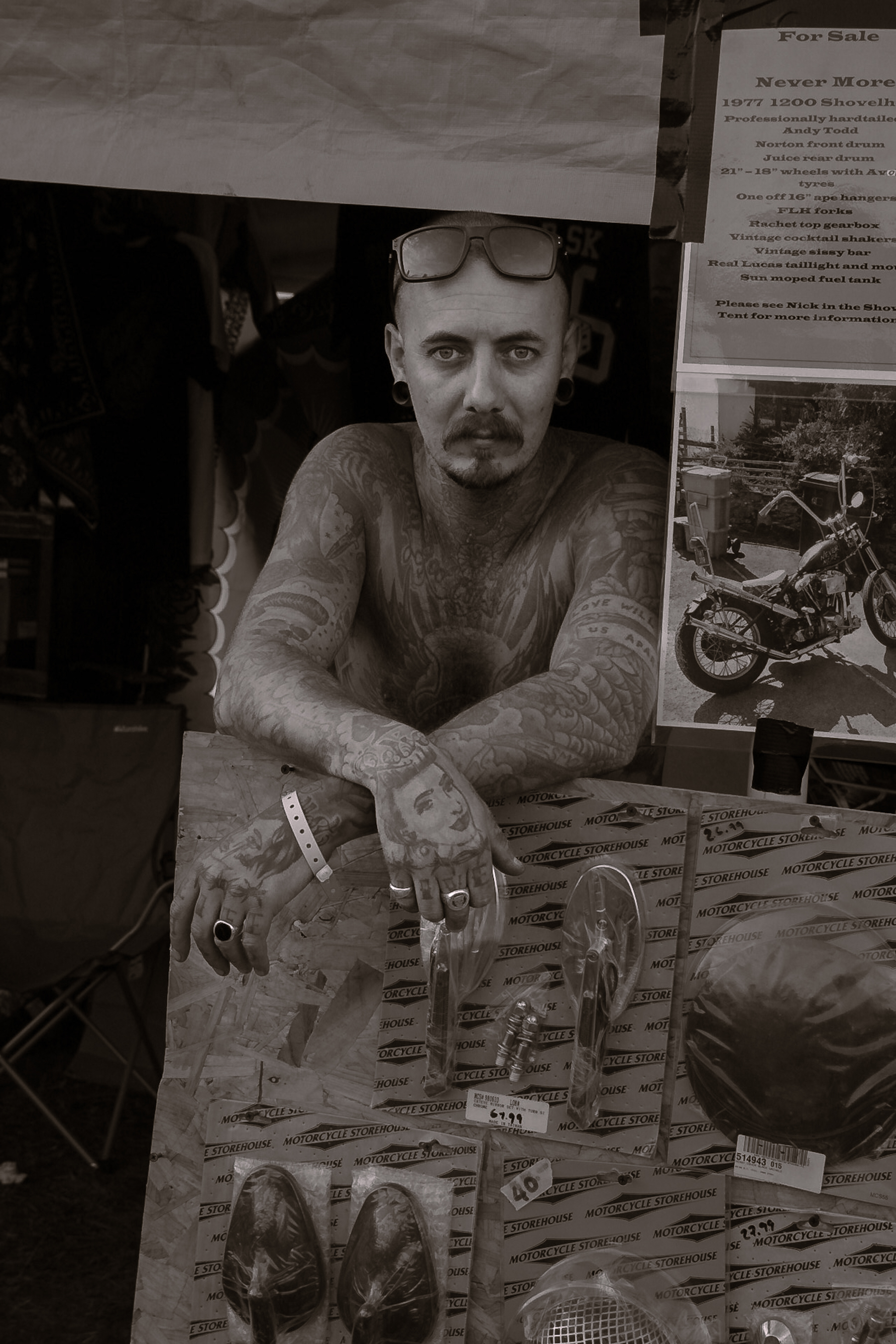 Monochrome portrait of a tattooed man with sunglasses on his head, leaning over a display of vintage motorcycle parts indoors.