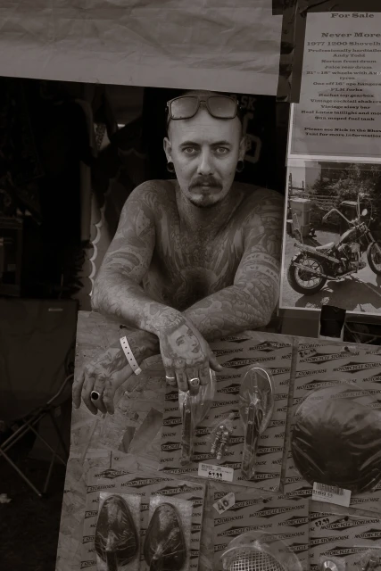 Monochrome portrait of a tattooed man with sunglasses on his head, leaning over a display of vintage motorcycle parts indoors.