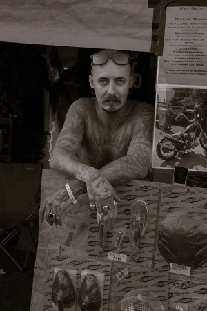 Monochrome portrait of a tattooed man with sunglasses on his head, leaning over a display of vintage motorcycle parts indoors.