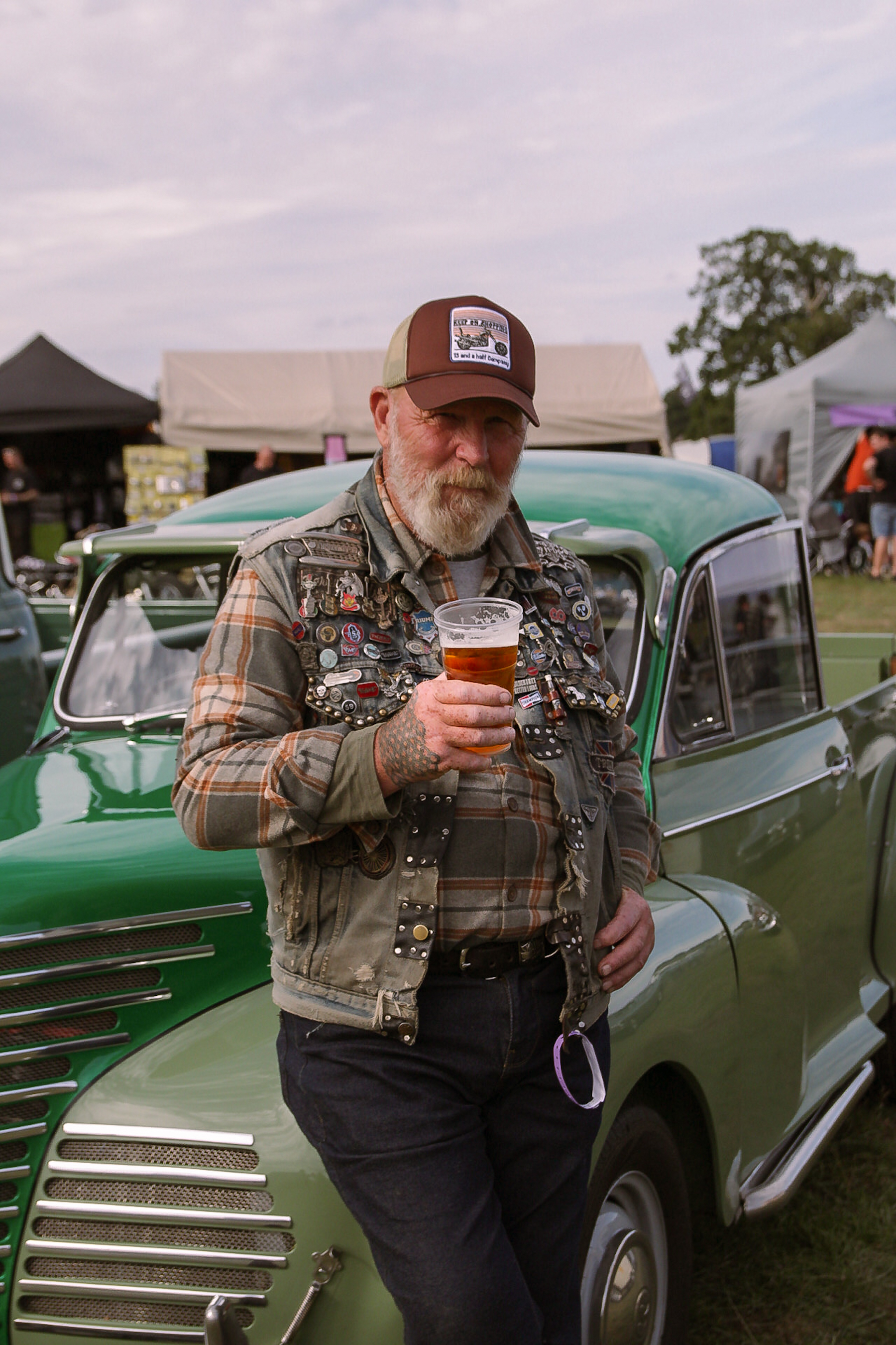 Older adult male wearing a hat with a beer in front of a green vintage car at an outdoor event on a cloudy day.