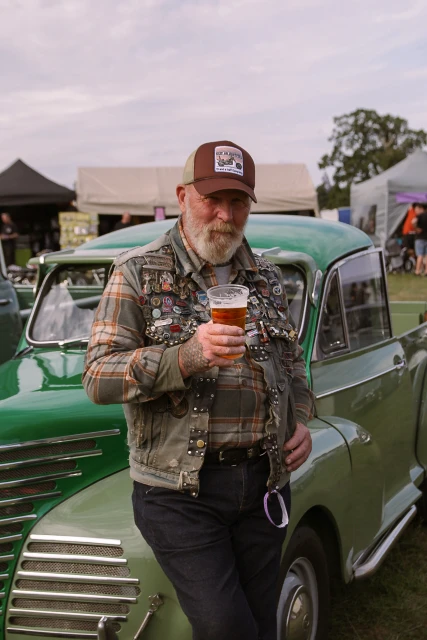 Older adult male wearing a hat with a beer in front of a green vintage car at an outdoor event on a cloudy day.