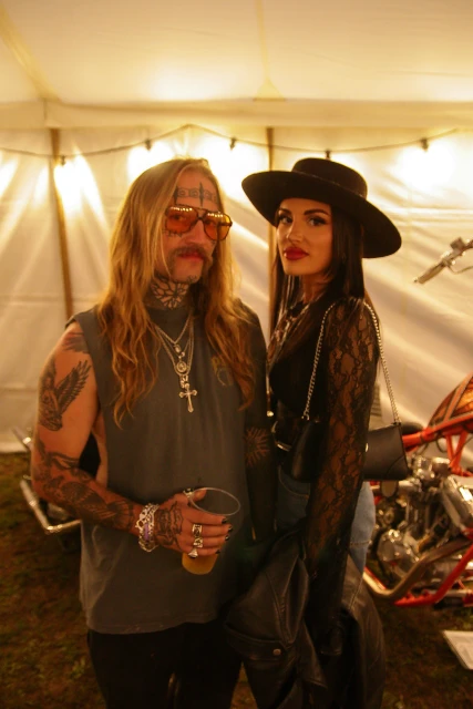 Couple posing outdoors in a tent with a custom motorcycle; the man has long hair and tattoos, and the woman wears a black hat and lace top.