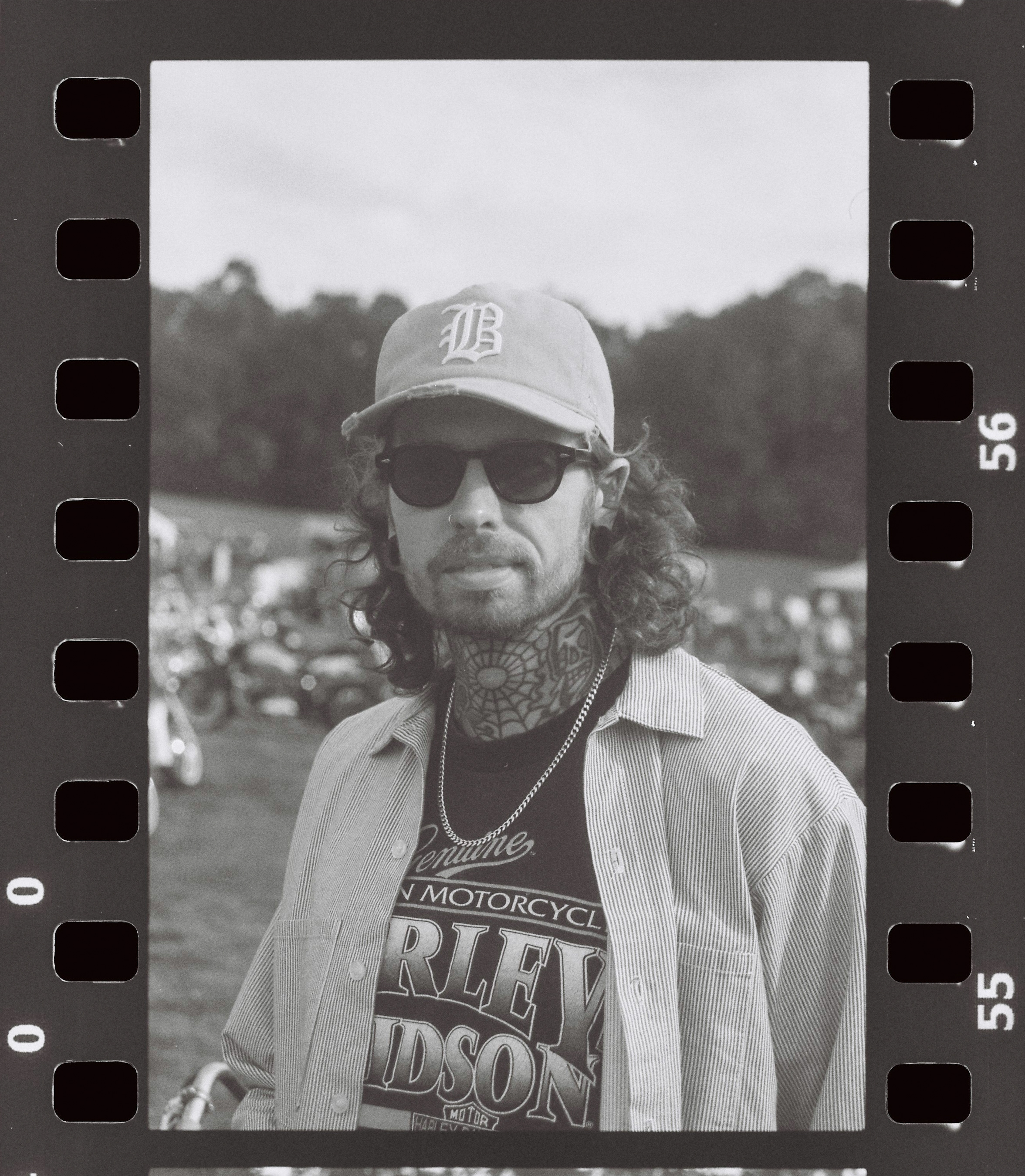 Monochrome portrait of a man with tattoos, wearing sunglasses and a baseball cap, posing in front of a blurred outdoor scene.