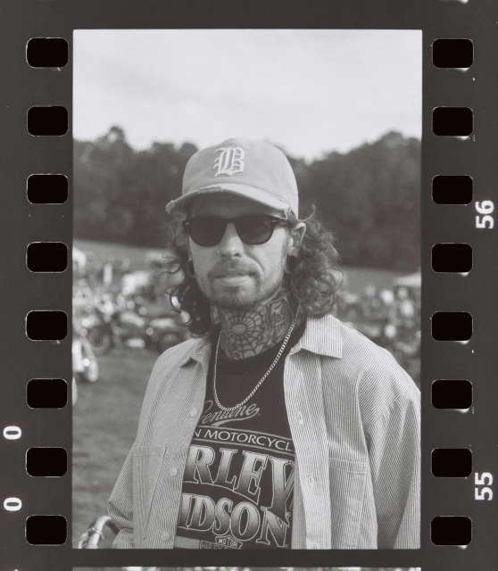 Monochrome portrait of a man with tattoos, wearing sunglasses and a baseball cap, posing in front of a blurred outdoor scene.